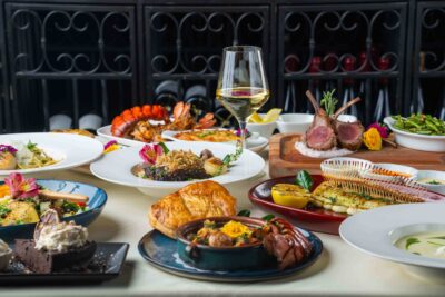 A luxurious spread of French Mediterranean dishes including escargot, puff pastry, lobster tails, grilled fish, lamb chops, and colorful cocktails and wine.