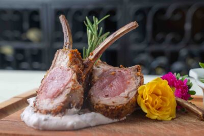 Two perfectly cooked rack of lamb chops served on a wooden board with vibrant edible flowers and a side of creamy sauce.