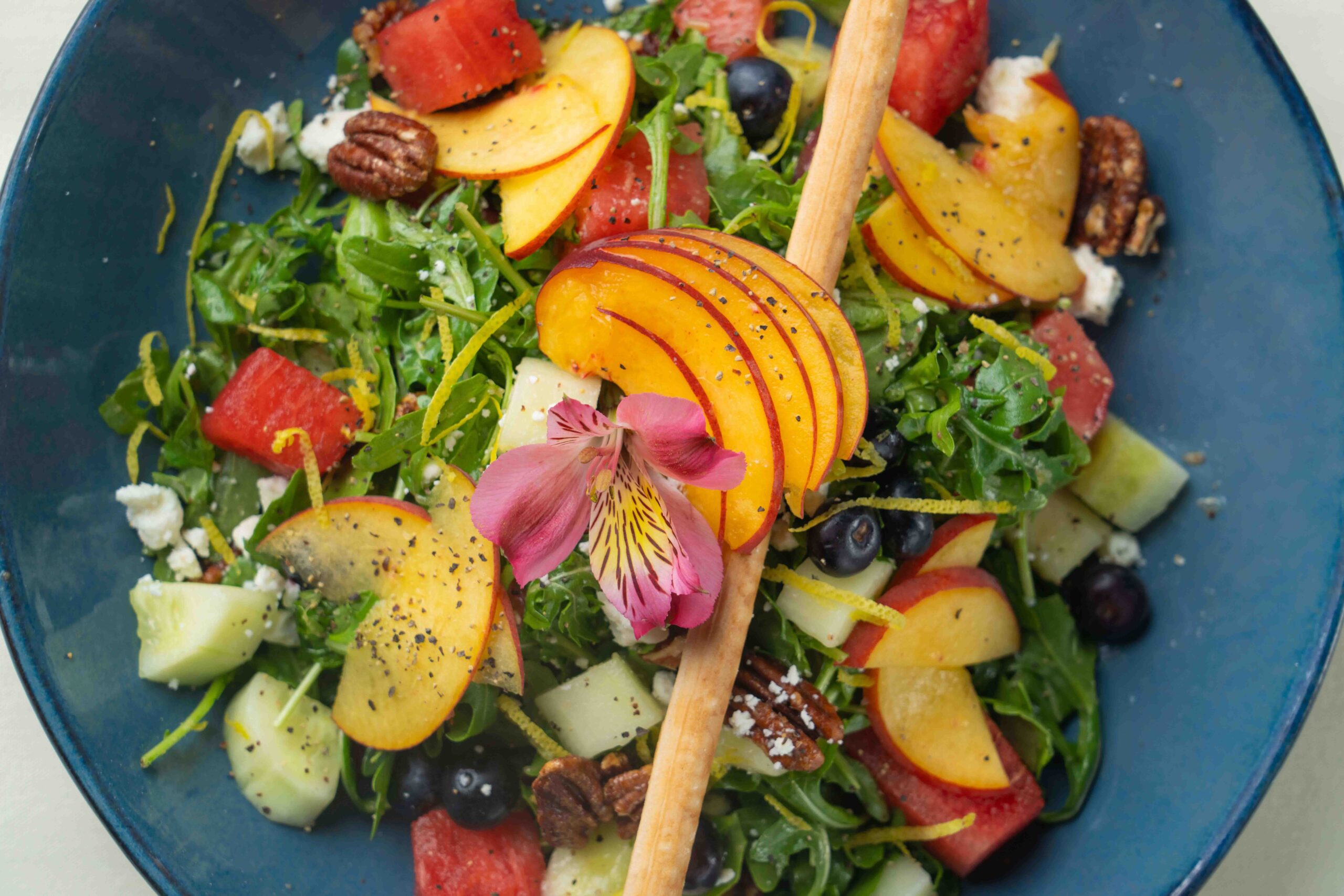 A colorful salad with fresh peaches, watermelon, cucumber, arugula, blueberries, pecans, and edible flowers on a blue plate.